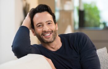 Portrait of a smiling happy young man resting while sitting on sofa. Handsome guy looking at camera. Young latin man in casual relaxing at home. Man relaxing on sofa with arm behind the head.