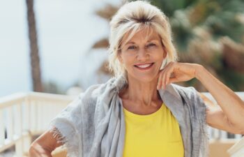 Stylish blond woman sitting on a balcony with a shawl over her shoulders in the sunshine smiling at the camera