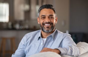 Portrait of happy mid adult man sitting on sofa at home. Handsome latin man in casual relaxing on couch and smiling. Cheerful indian guy looking at camera.
