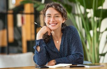 Portrait of smiling woman wearing spectacles while sitting at desk. Business woman taking notes in diary and looking at camera. University girl with eyeglasses sitting on table at library.