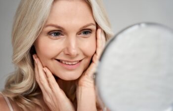 Happy 50s middle aged woman model touching face skin looking in mirror. Smiling mature older lady pampering, enjoying healthy skin care, aging beauty, skincare treatment cosmetic products concept.
