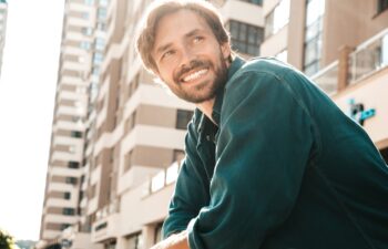 Portrait of handsome smiling stylish hipster lambersexual model.Man dressed in green shirt. Fashion male posing in the street at skyscraper background
