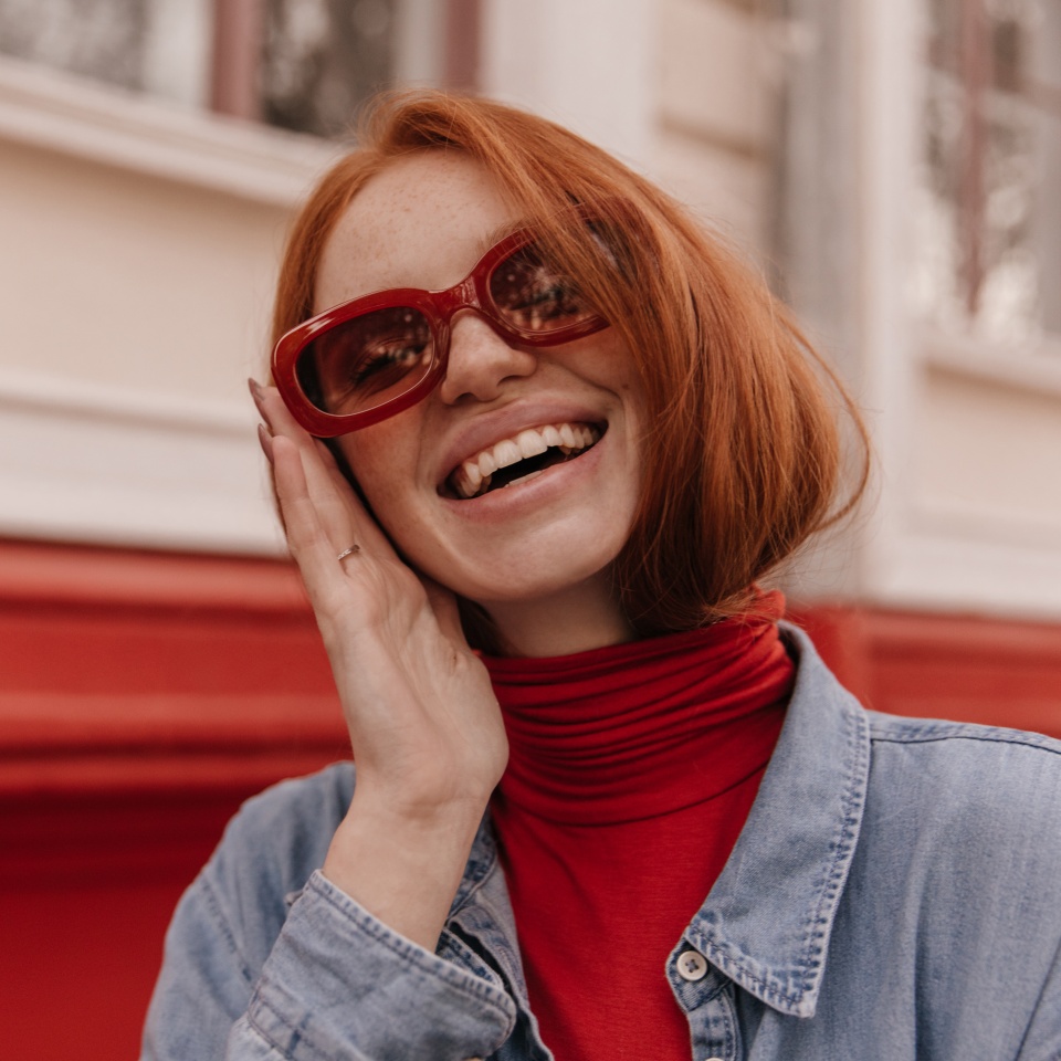 Close-up portrait of young beautiful lady posing outdoors. Pretty red-haired girl wearing turtleneck and blue shirt, adjusting trendy sunglasses and smiling against bright wall background