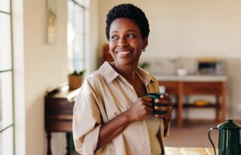 Happy Brazilian woman looking outside the window in her home kitchen, smiling and holding an enamel mug of plain coffee. Mature, afro-haired woman standing next to a breakfast table.