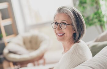 Photo of charming good mood elderly lady wear white cardigan spectacles smiling indoors apartment room