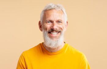 Closeup portrait handsome smiling 50 years old man with stylish gray hair wearing casual orange t shirt, looking at camera isolated on background. Health care, dental treatment concept