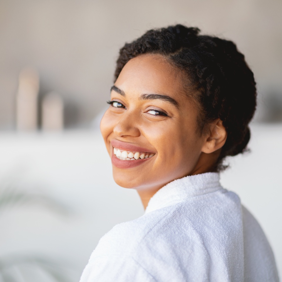 African American woman with curly hair smiles brightly while wearing a cozy spa robe. She seems relaxed and content in a calm and soothing setting, radiating happiness and tranquility, copy space