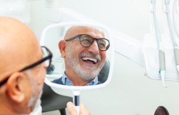 Happy senior patient admiring his perfect smile after a dental implant treatment, holding a mirror in his hand at the dental clinic